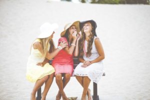 Three pretty girls sitting on a bench and chatting. They are wearing big summer hats.