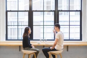 A guy and a girl drinking coffee next to a large window and chatting away.